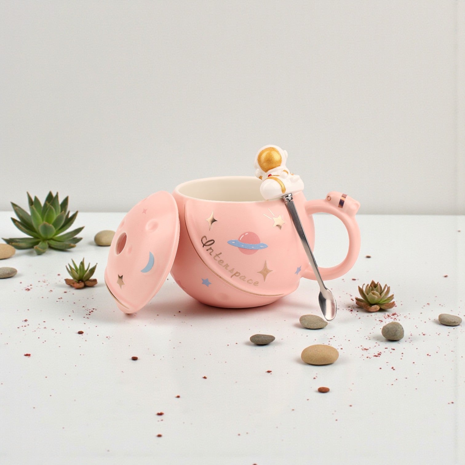 Pink ceramic mug with space-themed illustrations, matching lid with gold astronaut finial, and metal spoon