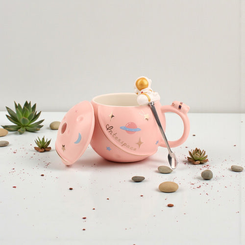 Pink ceramic mug with space-themed illustrations, matching lid with gold astronaut finial, and metal spoon