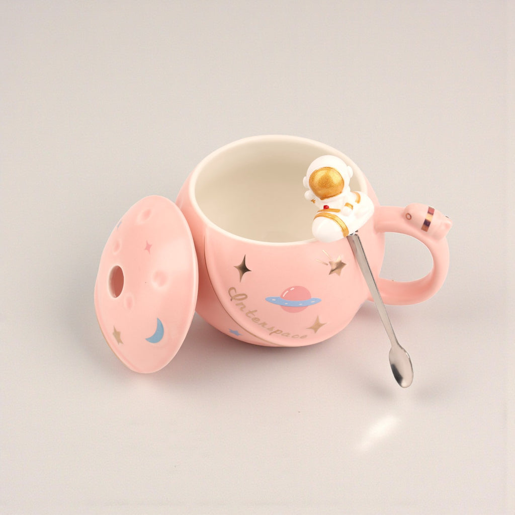 Pink ceramic mug with space-themed design, astronaut figurine handle, and matching lid with moon and planet illustrations