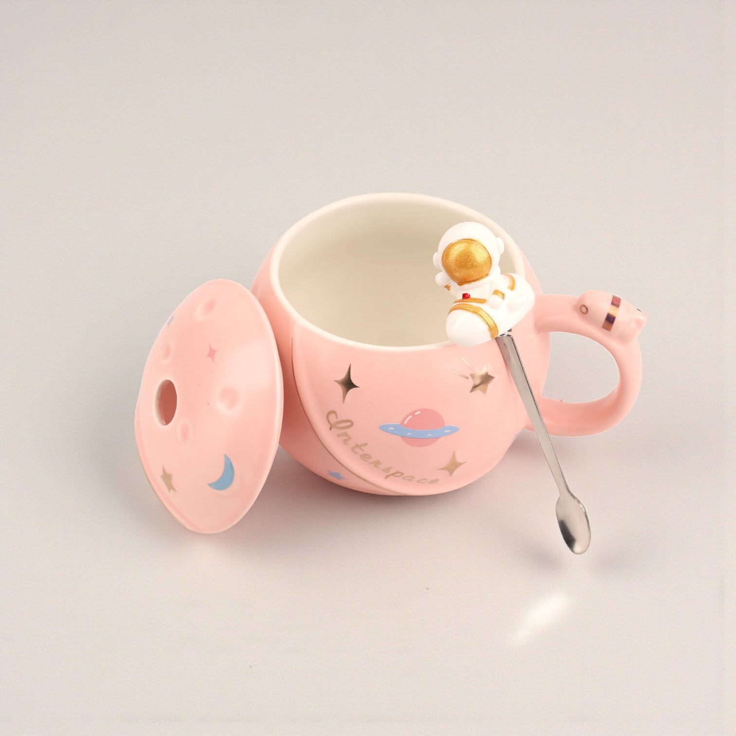 Pink ceramic mug with space-themed design, astronaut figurine handle, and matching lid with moon and planet illustrations