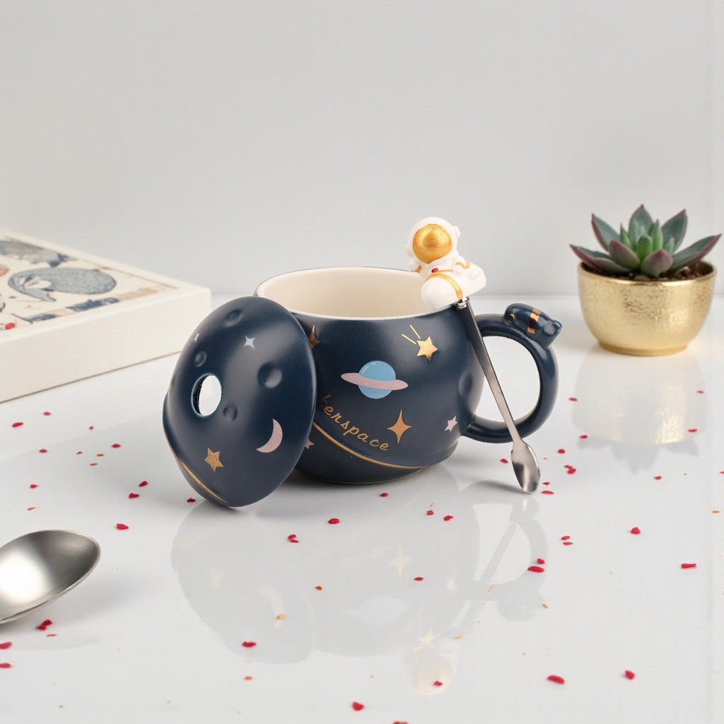 Dark navy ceramic mug with gold stars, moon, and planet design, matching lid with astronaut spoon and gold spaceman finial