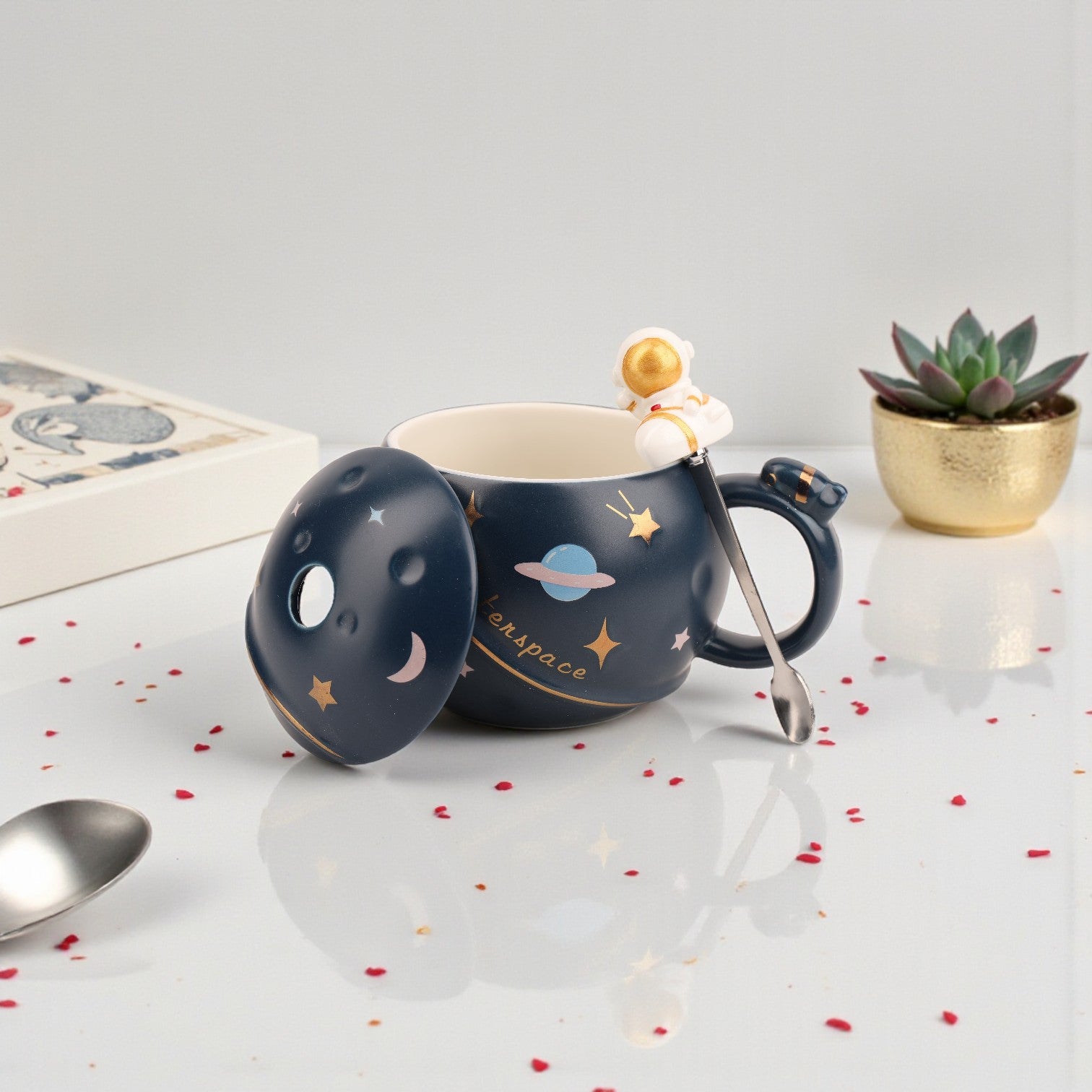 Dark navy ceramic mug with gold stars, moon, and planet design, matching lid with astronaut spoon and gold spaceman finial