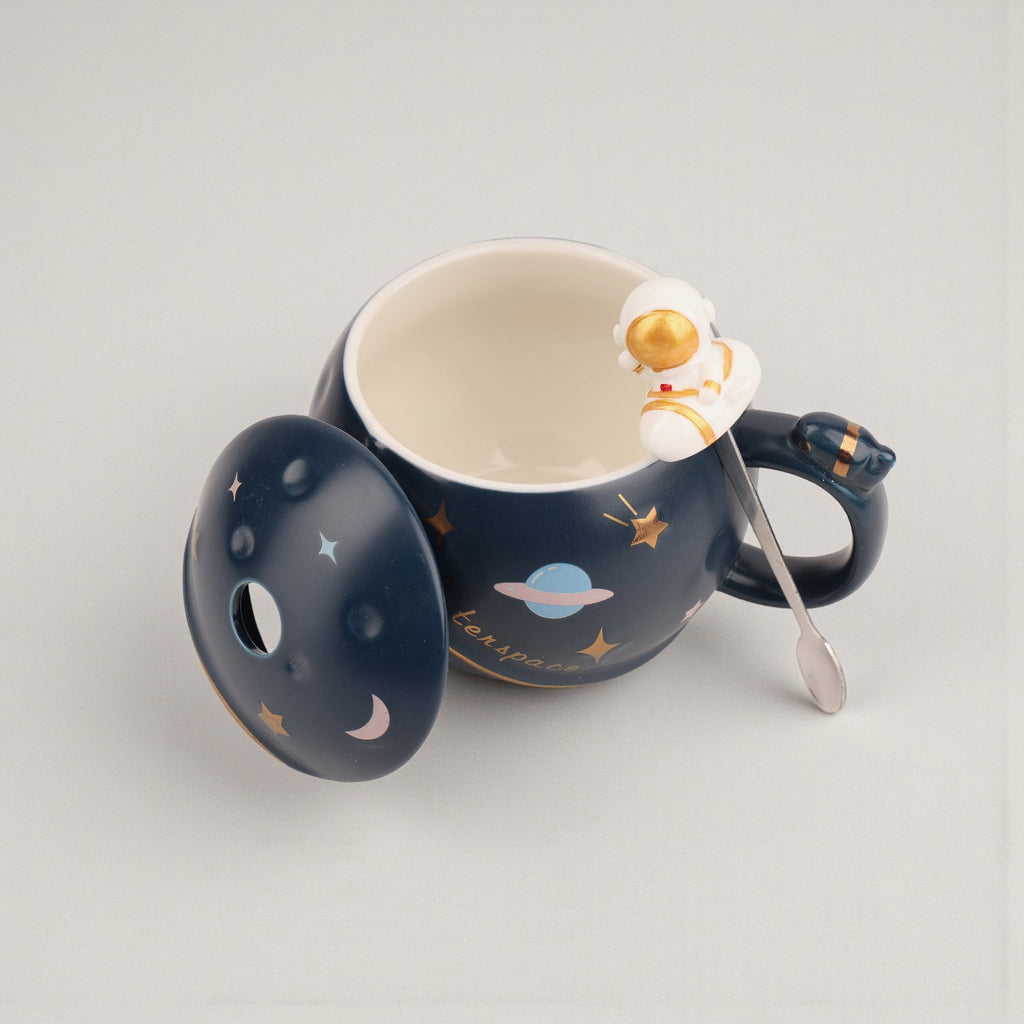 Dark blue ceramic mug with gold stars, planet, and astronaut figurine on rim, cream interior with matching spoon