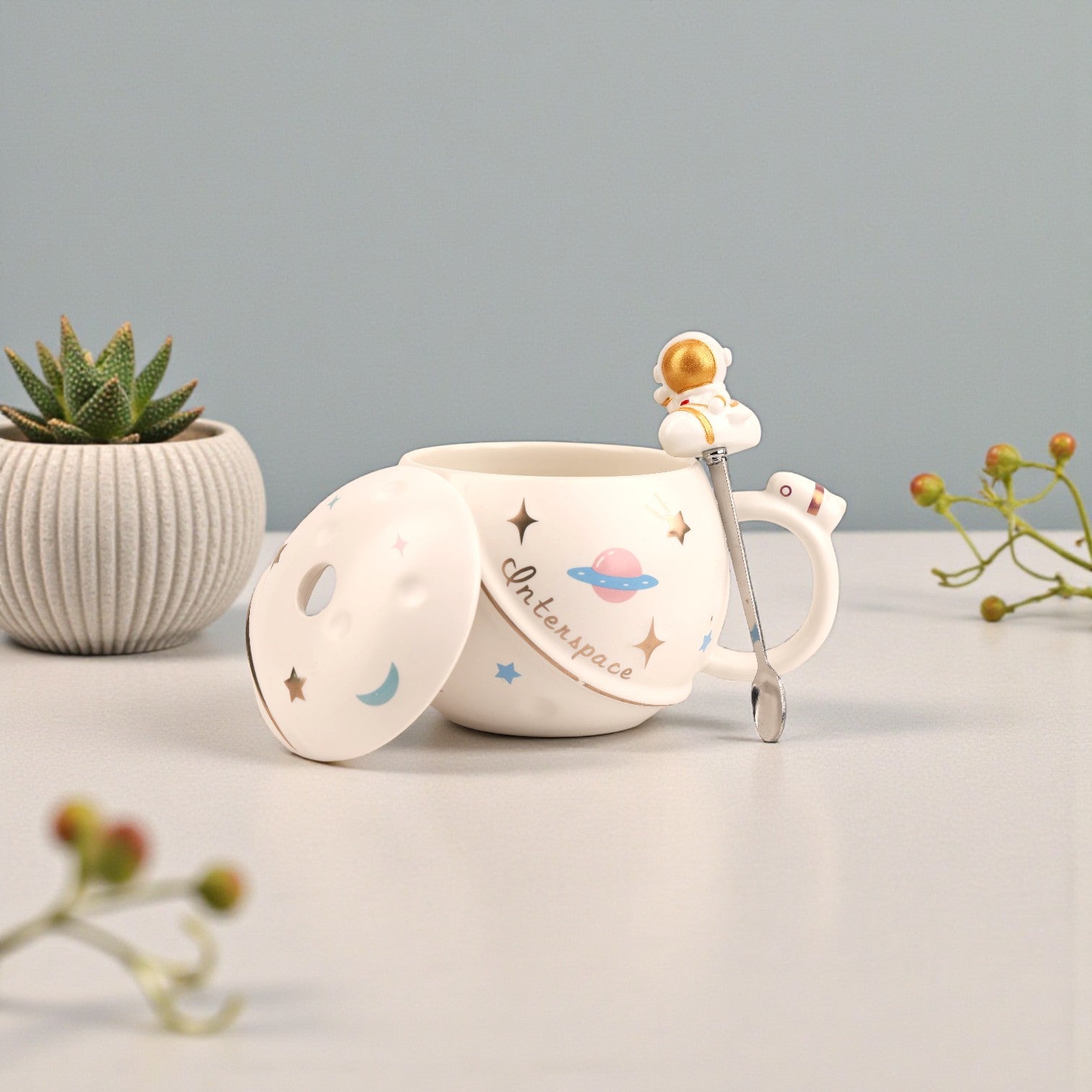 White ceramic mug with space-themed illustrations, matching lid, and astronaut spoon with gold helmet