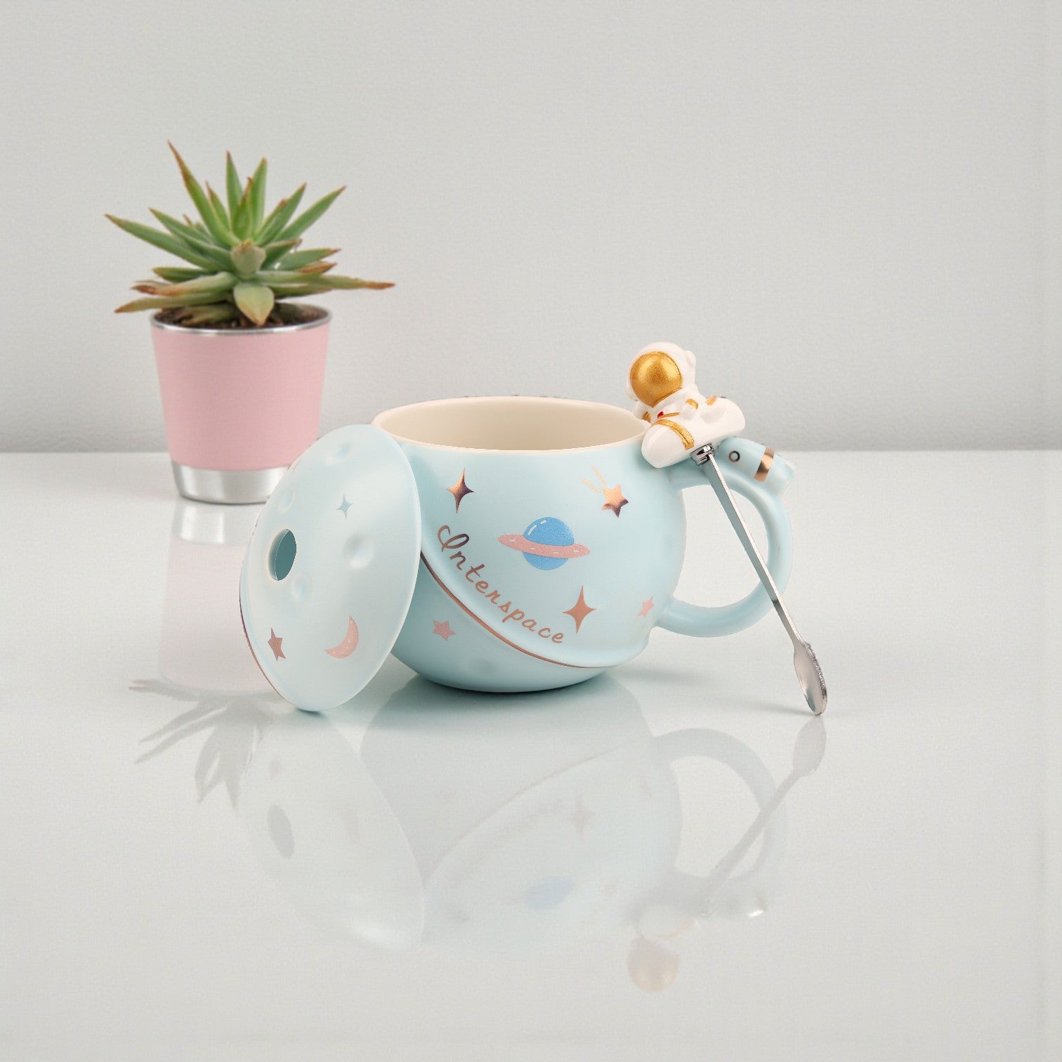 Light blue ceramic mug with space-themed illustrations, matching lid, astronaut spoon, and gold planet charm accent
