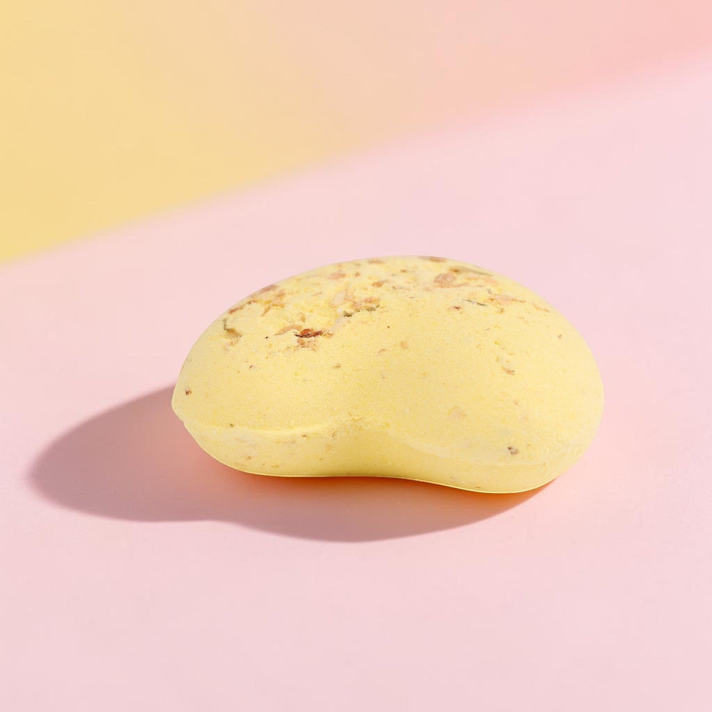 Pale yellow oval bath bomb with speckled texture on pink gradient background