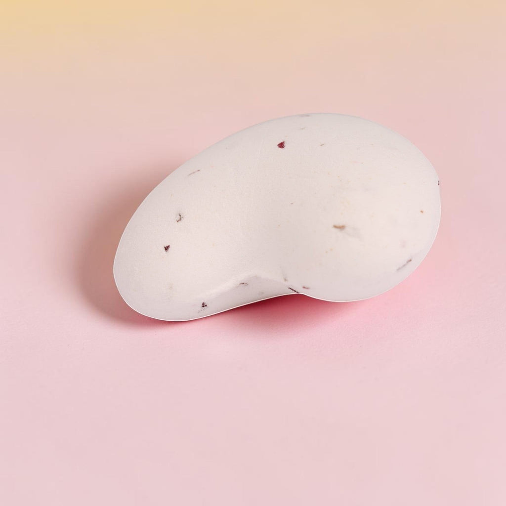 White oval bath bomb with rose petals and botanical flecks on pink background